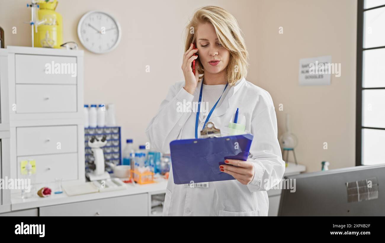 Blonde woman in lab coat talks on phone holding clipboard in laboratory Stock Photo