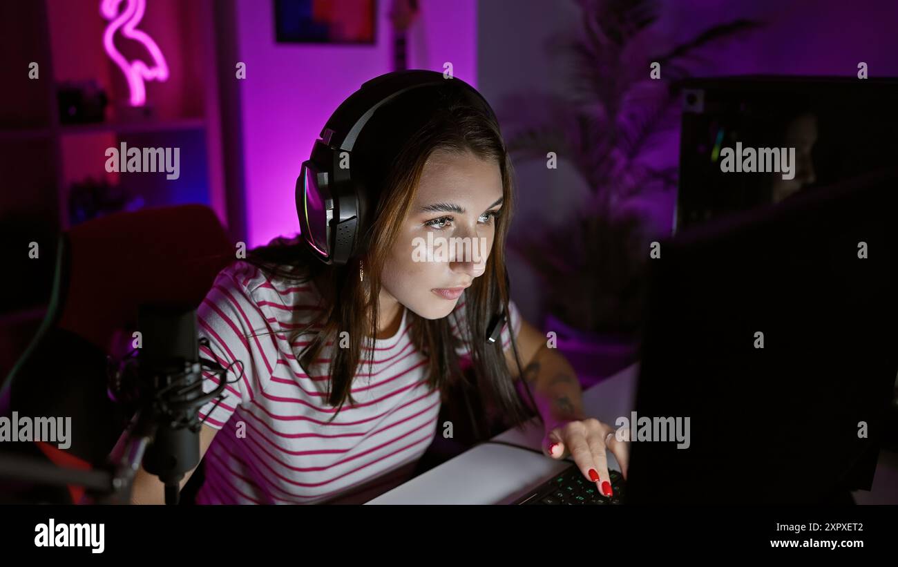 Focused young woman with headphones using computer in a dark gaming room, illuminated by neon light. Stock Photo