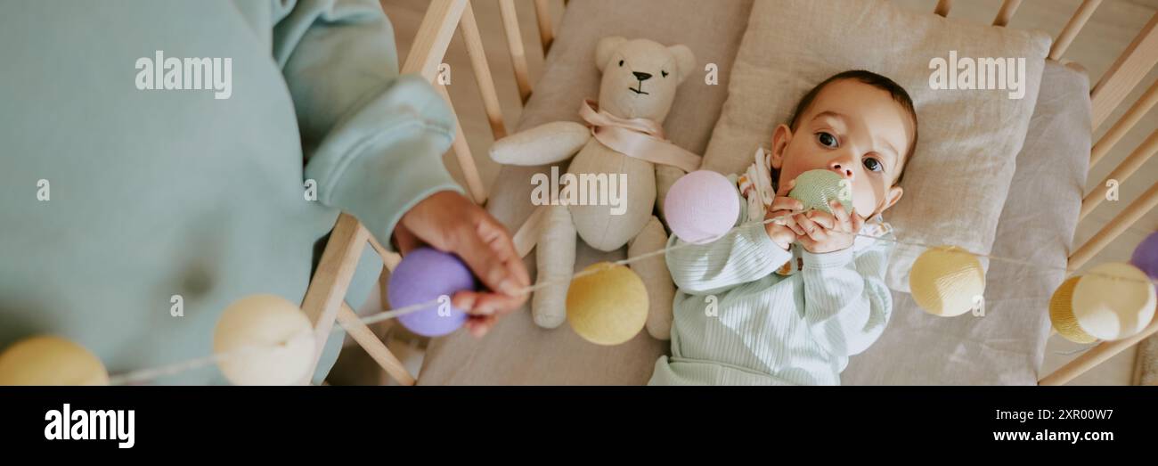 Website banner of baby boy playing with multicolored rattle and biting it white unrecognizable female hands adjusting rattle Stock Photo