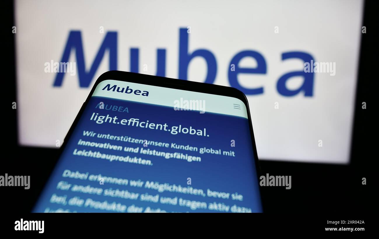 Mobile phone with website of German automotive company Muhr und Bender KG (Mubea) in front of business logo. Focus on top-left of phone display. Stock Photo