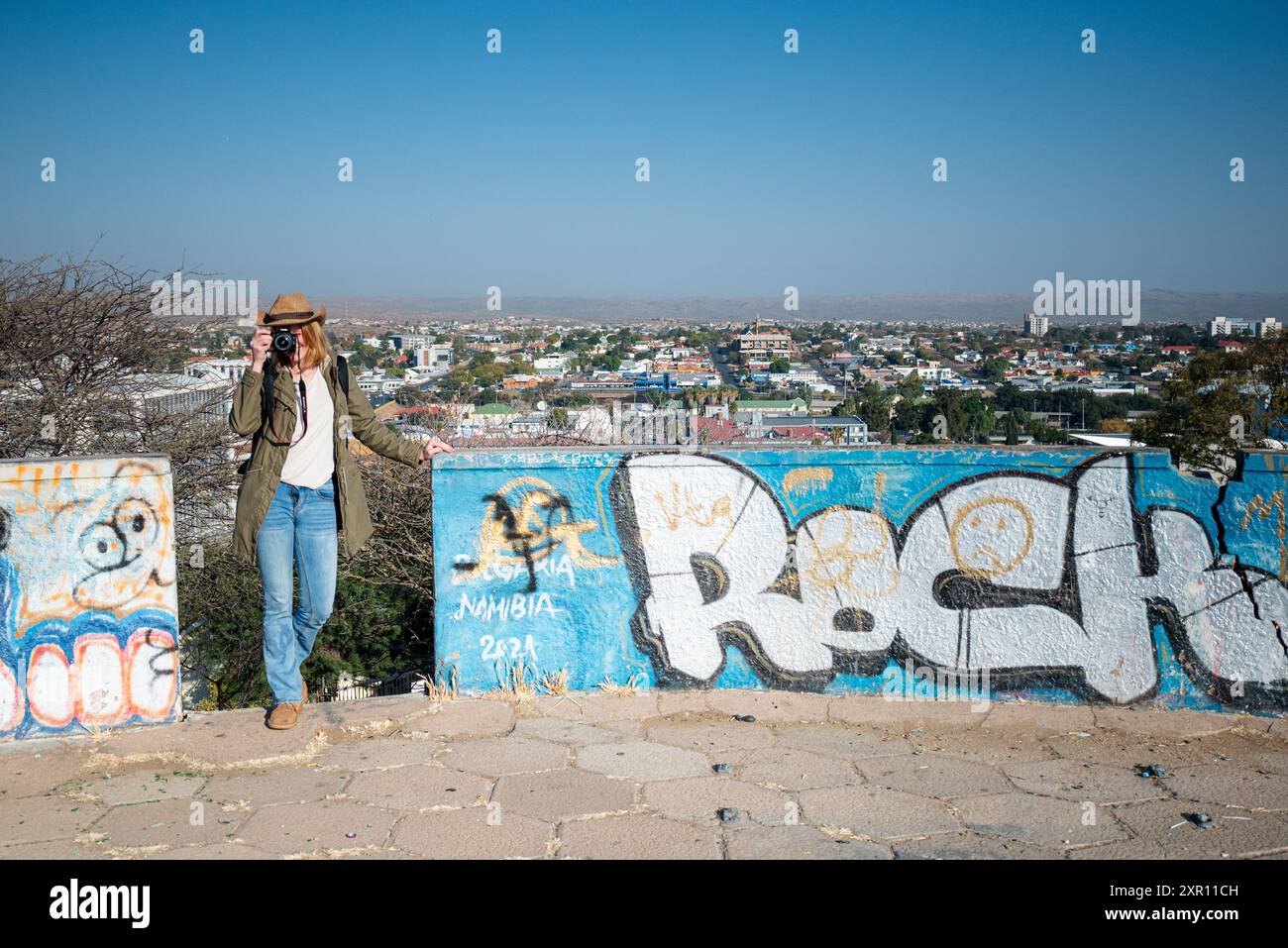 A tourist taking a photo with a camera in front of graffiti art on a wall overlooking a cityscape in Windhoek, Namibia. Stock Photo