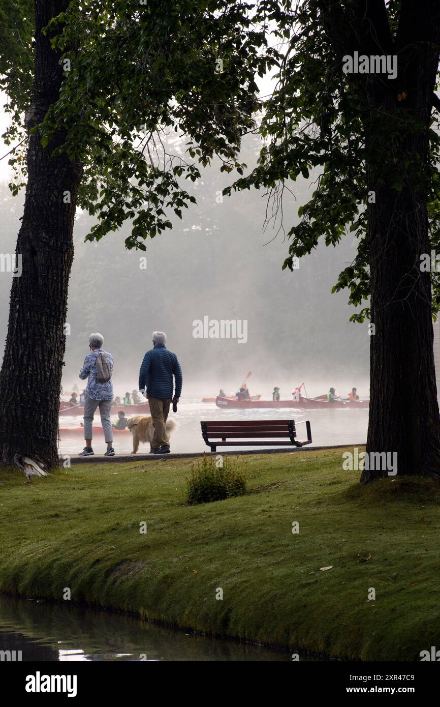 An older couple walking their dog while canoeists and kayaks paddle in the distance. Bowness Park, Calgary, Alberta Canada Stock Photo