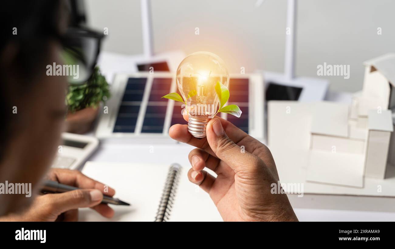 Close-up at a light bulb, An engineer sits holding a light bulb and model of wind turbine. with solar panels and model house on the table. To design u Stock Photo