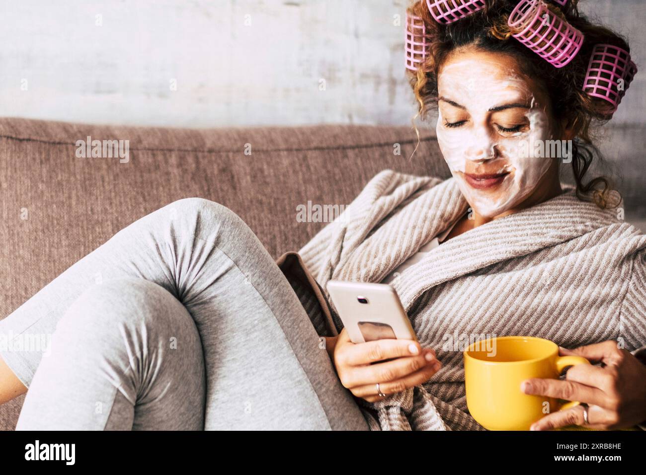 One young woman have relax leisure activity alone at home with care treatment skin face cream product and curlers on hair, Beauty caring on sofa and using phone connection. Female lifestyle routine Stock Photo