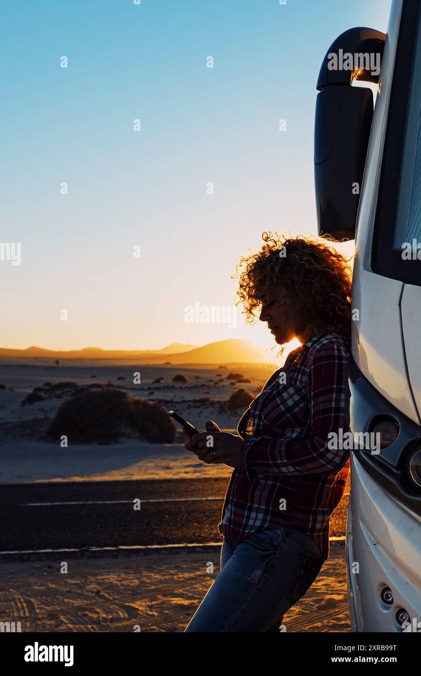 People on travel with modern camper. Woman using mobile phone roaming against big motorhome. Concept of modern lifestyle and travelers. Vacation and vanlife sending message with cellular. Everywhere internet connection Stock Photo