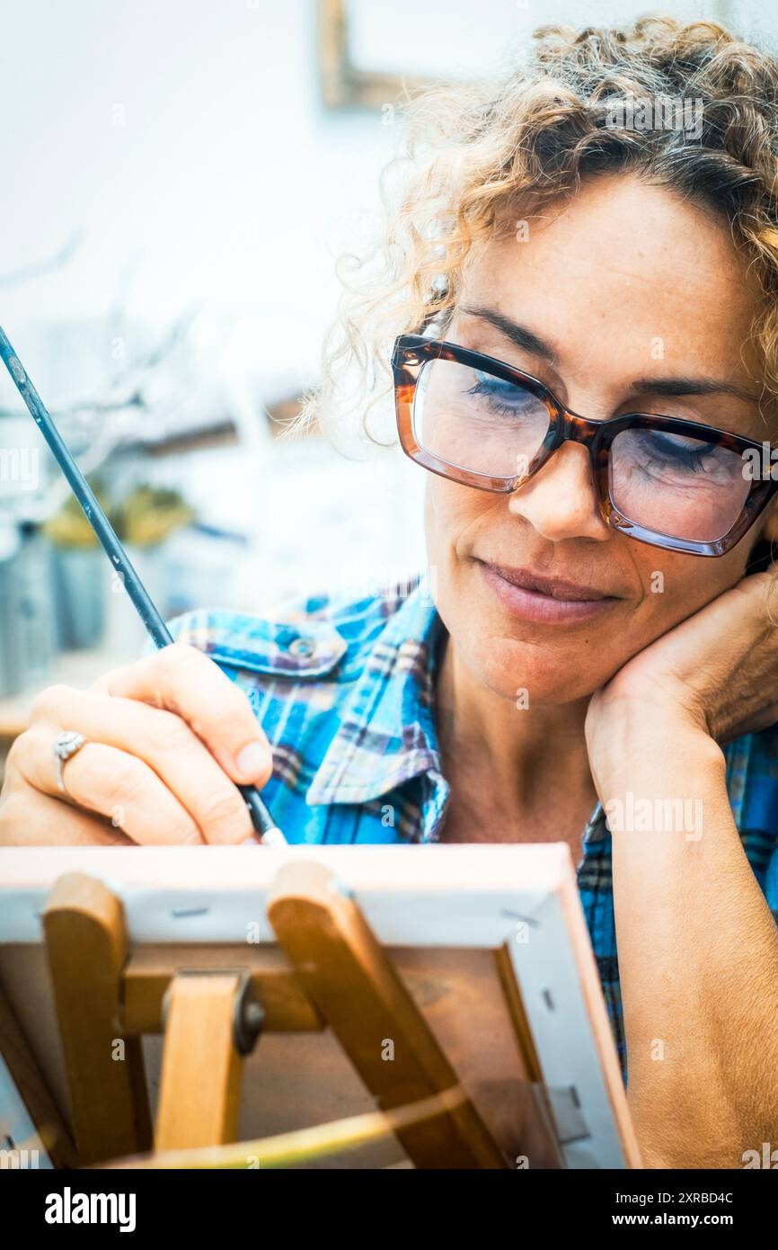 Picture of serious concentrated adult Caucasian female artist sitting at desk with painting accessories, looking the canvas and paint enjoying hobby and indoor leisure activity alone. Mature lady home Stock Photo