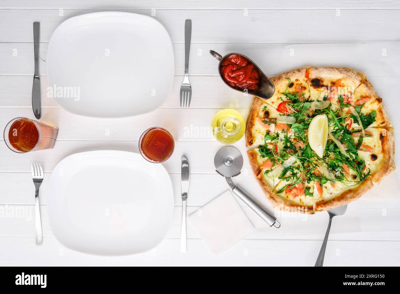 Freshly cooked pizza, cutlery, sauces and drinks for two people on white wood table Stock Photo