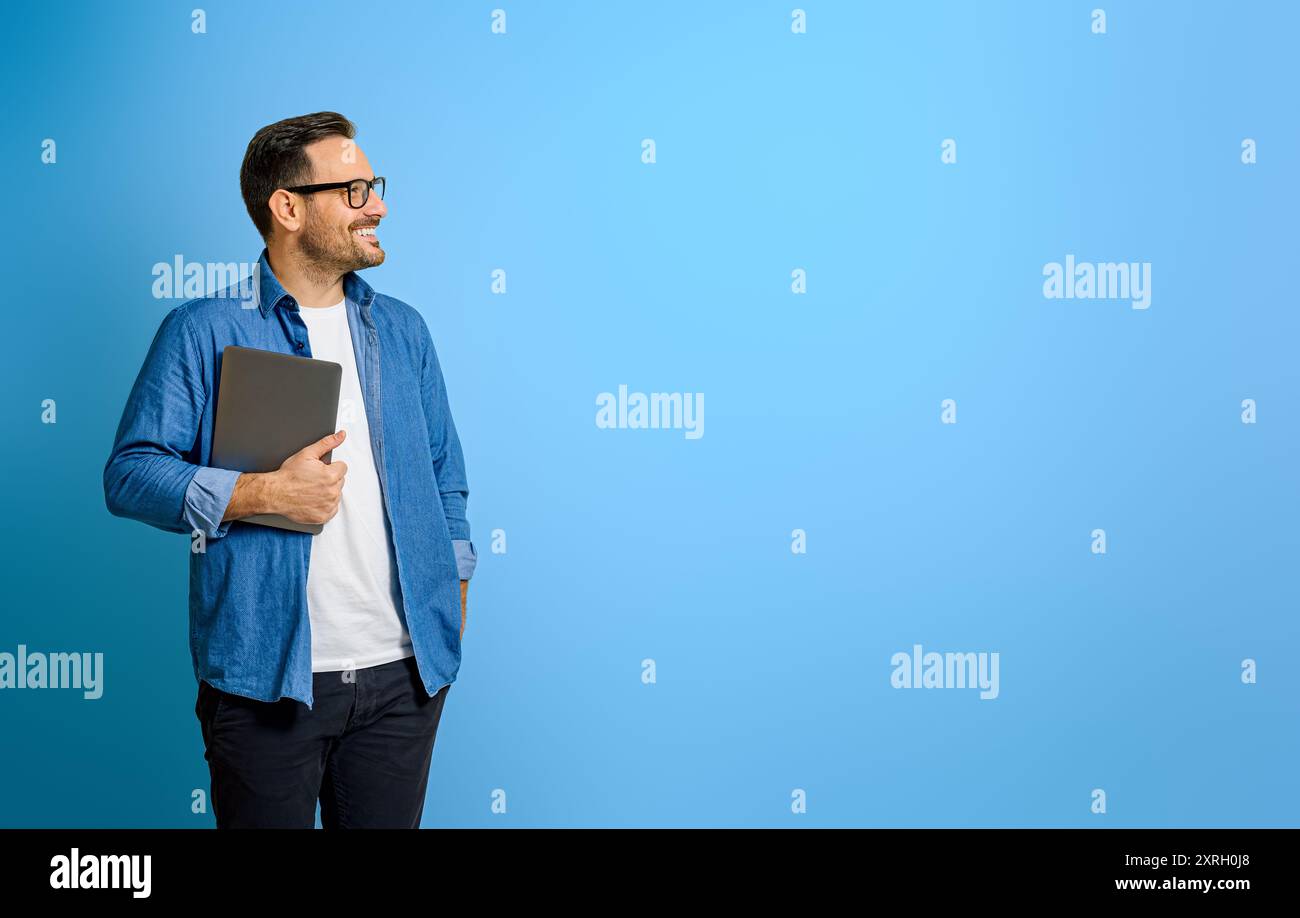 Thoughtful smiling businessman holding digital tablet and looking away against blue background Stock Photo
