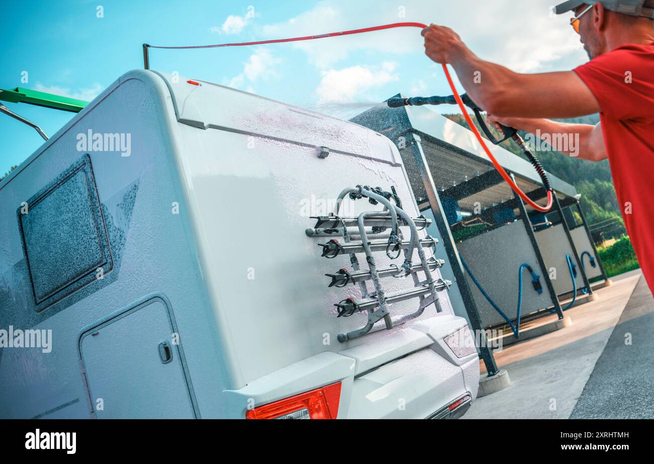 A man sprays water on the back of a camper van, using a hose at a car wash station under a clear blue sky. Stock Photo