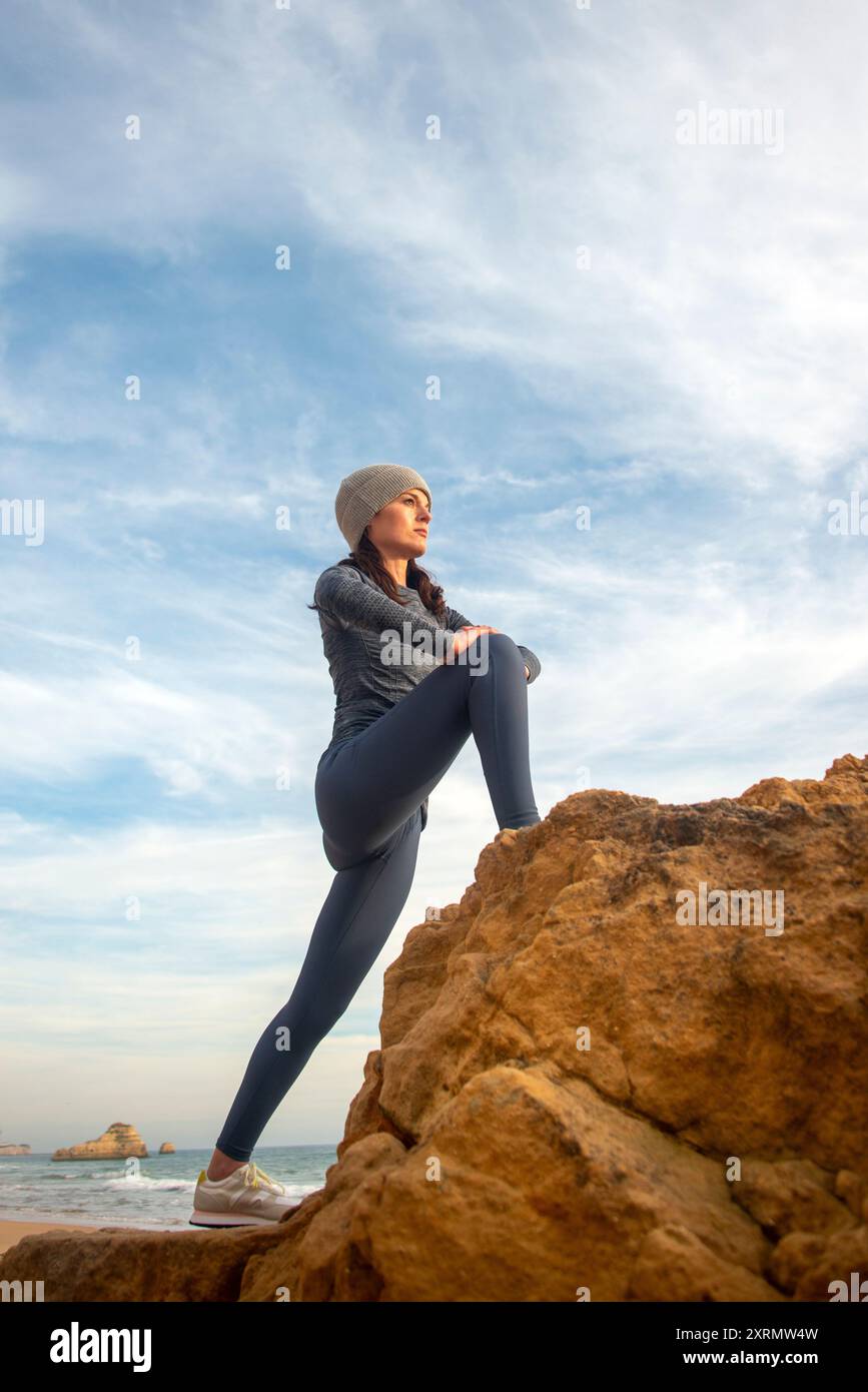 Sporty woman jogger doing leg stretching exercise on rocks by the sea. Stock Photo