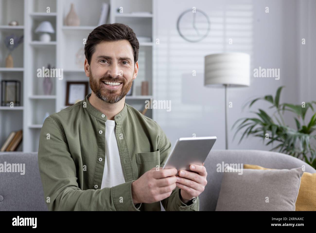 Man sitting on sofa smiling while using tablet at home. Relaxed and cheerful expression, wearing casual clothing, and engaging with device. Bright and cozy living room environment Stock Photo