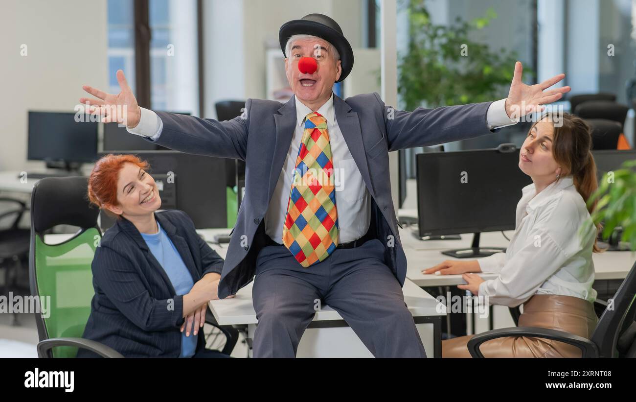 An elderly Caucasian man in a clown costume amuses two Caucasian women in the office.  Stock Photo