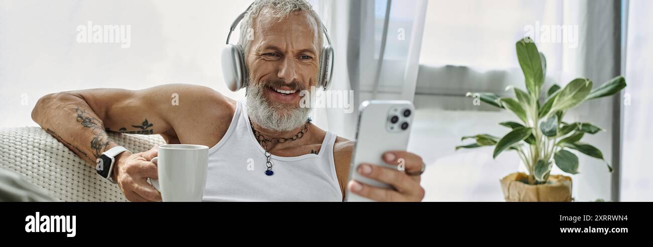 A mature man with grey beard and tattoos is smiling and relaxing on a couch at home. He is wearing headphones and looking at his phone. Stock Photo