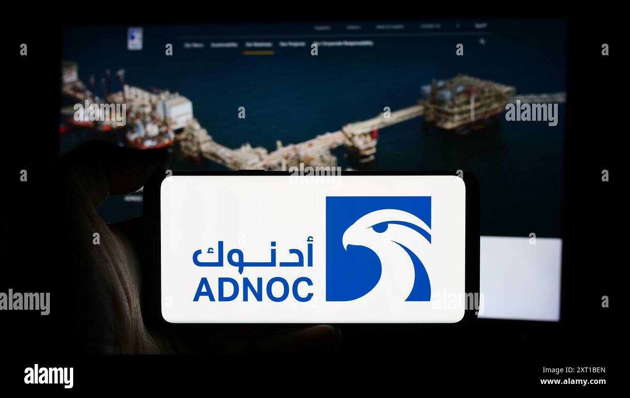 Person holding cellphone with logo of oil business Abu Dhabi National Oil Company (ADNOC) in front of business webpage. Focus on phone display. Stock Photo