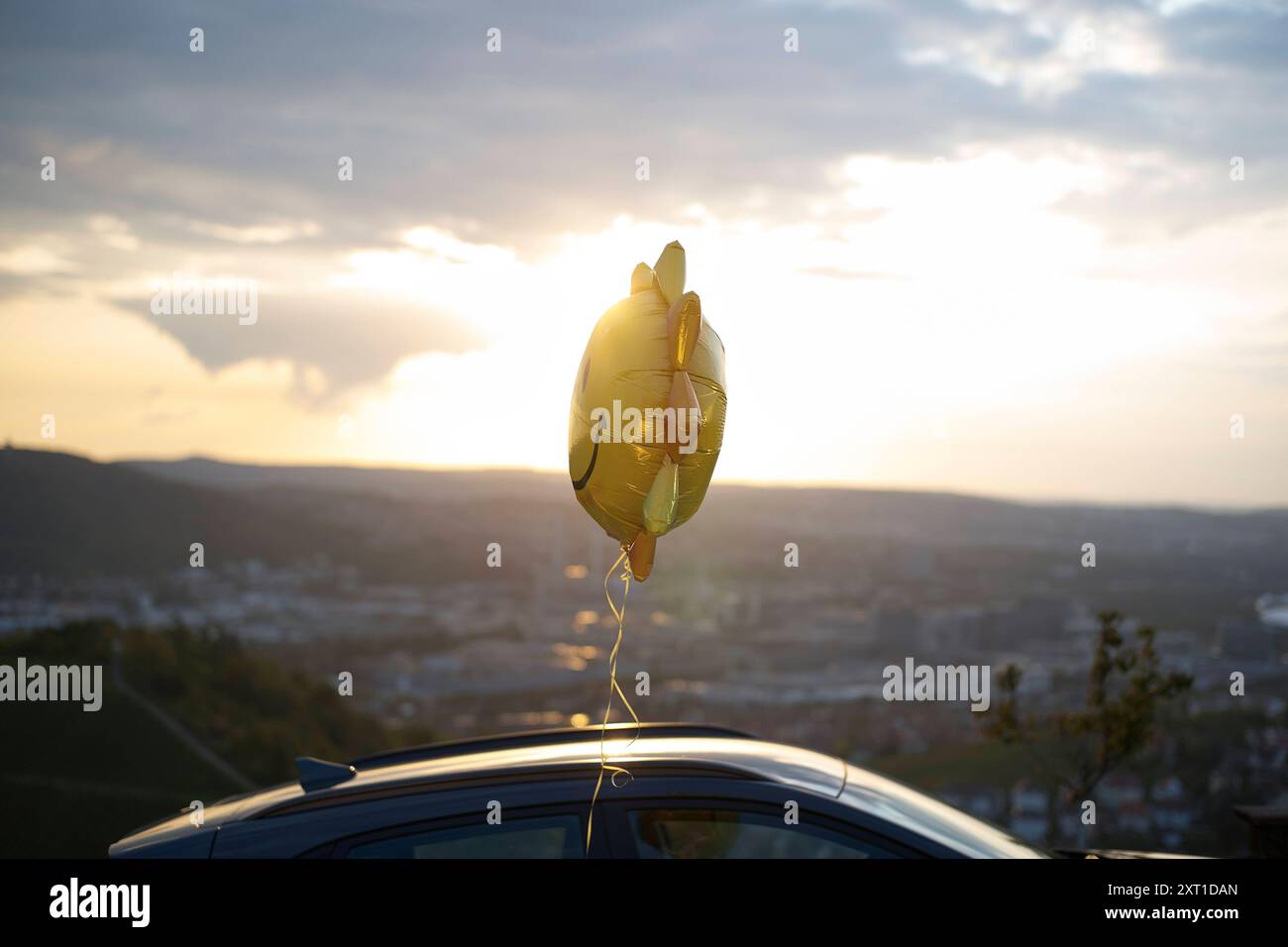 Golden star-shaped balloons tied to a car roof with a scenic sunset and landscape in the background. bola02373 Copyright: xConnectxImagesx Stock Photo