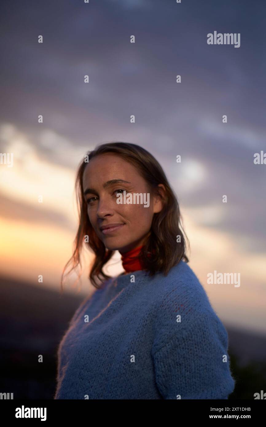 Smiling woman in a blue sweater with a red scarf standing against a sunset sky background. bola02413 Copyright: xConnectxImagesx Stock Photo