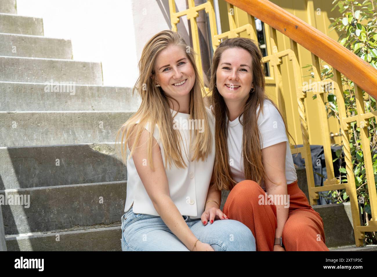 Two smiling women sitting together on steps with a yellow handrail in the background. panc05408 Copyright: xConnectxImagesx Stock Photo