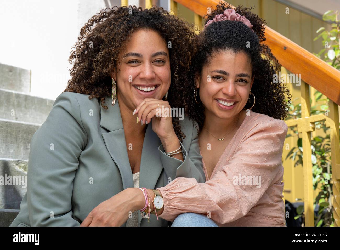 Two smiling women sitting together on steps, one in a green blazer, the other in a pink jacket, both appearing happy and relaxed. panc05401 Copyright: Stock Photo
