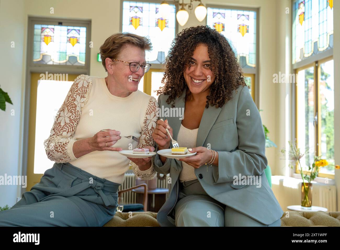 Two women smiling, enjoying tea and cake in a room with stained glass windows. panc05687 Copyright: xConnectxImagesx Stock Photo