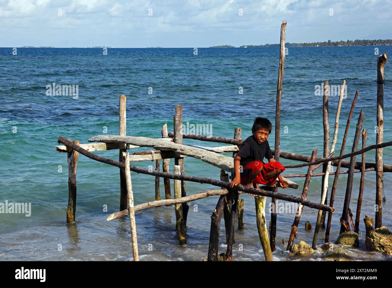 Kuna boy stting on rustic jetty made of wooden poles, sticks and beams, Carti Sugtup Island, San Blas Islands, Panama Stock Photo