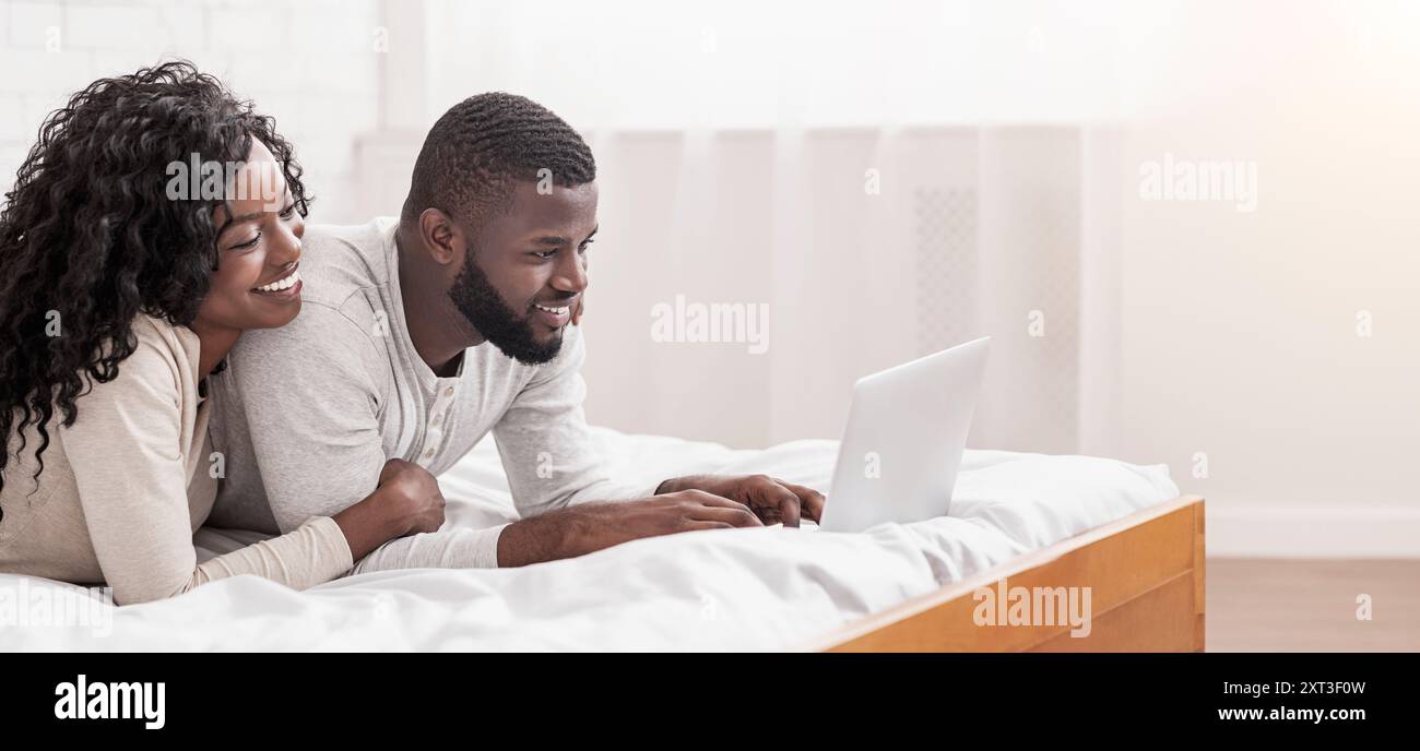 Cheerful afro spouses lying in bed using laptop together Stock Photo