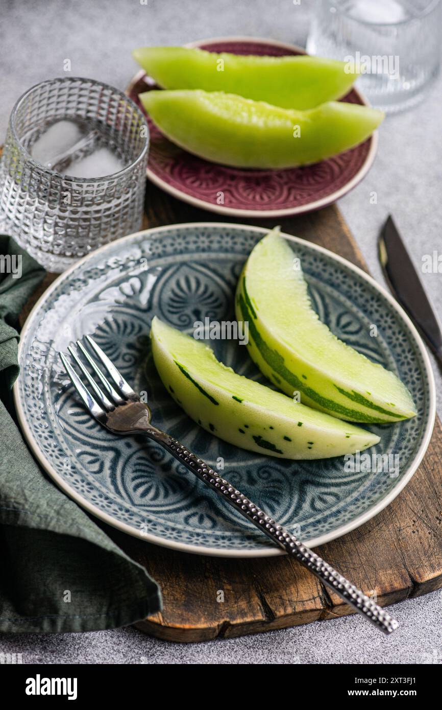 From above organic mini melon slices arranged on ornate ceramic plates, accompanied by a glass of water with ice and a fork on a textured tablecloth. Stock Photo