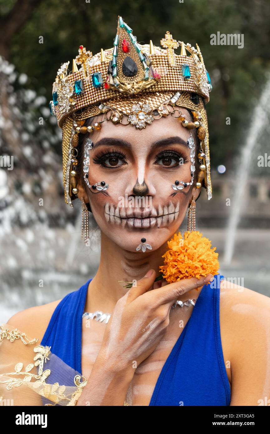 A woman in detailed makeup representing a Catrina, adorned with a sparkling crown and holding a marigold, celebrates Day of the Dead in Mexico Stock Photo