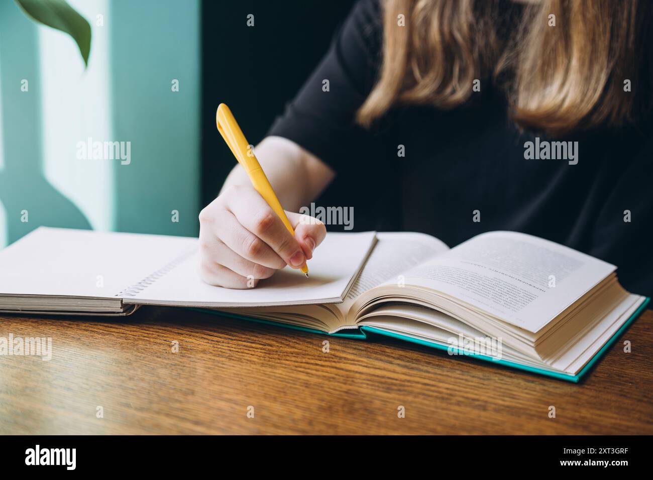 Cropped unrecognizable photo of a female student looking down while writing notes in a book, sitting at a wooden table, with a focus on her hands and Stock Photo