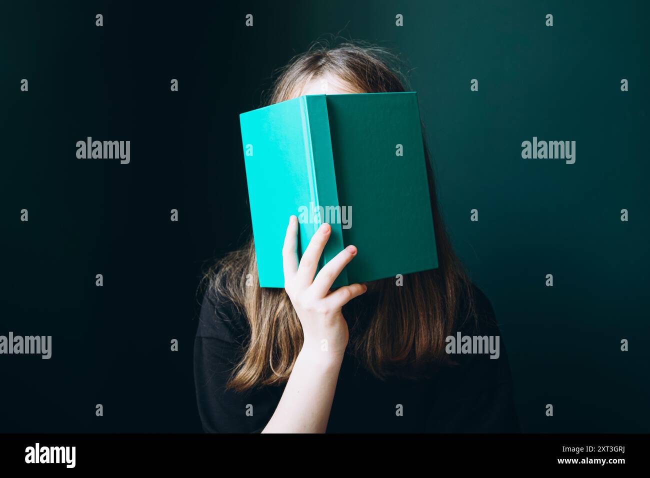 Cropped and unrecognizable young female student covering her face with a teal book against a dark background, suggesting anonymity and intrigue in an Stock Photo