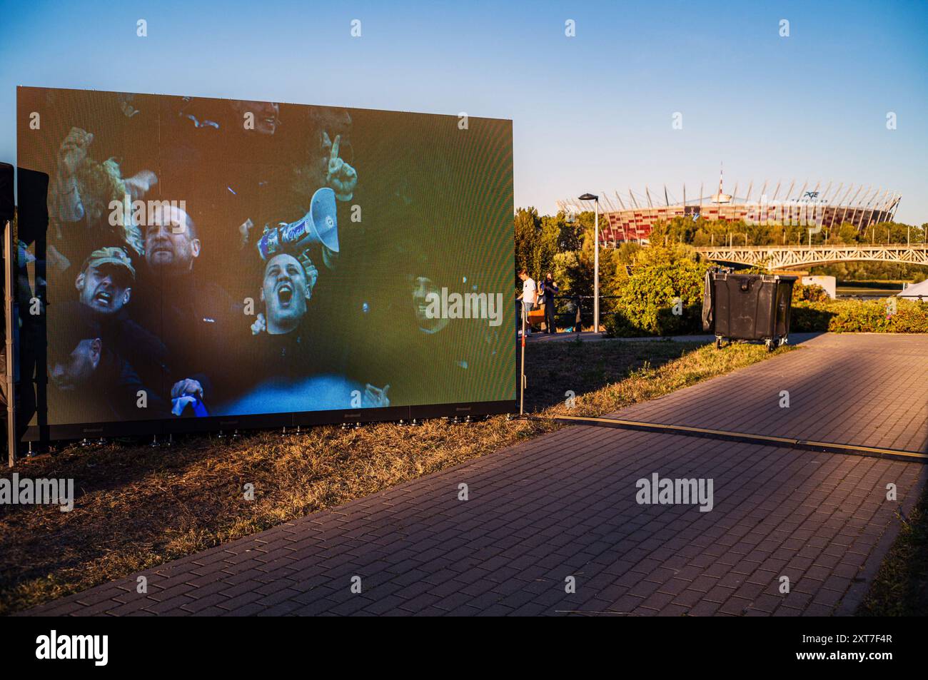 A screen shows images of football fans celebrating, while the venue of the Super Cup final looms in the background. Fans of Spanish Champions League winners, Real Madrid, and Italian UEFA Cup winners, Atalanta, gathered in Kahla Square in Warsaw, beside the Vistula river, for the UEFA Super Cup fan festival. In anticipation of the match the following day, visitors queue to make photos with the trophy, and take part in a number of football-related games and activities. The UEFA Super Cup takes place at PGE Narodowy in Warsaw on 14th August 2024 and is contested by the winners of the Champions L Stock Photo