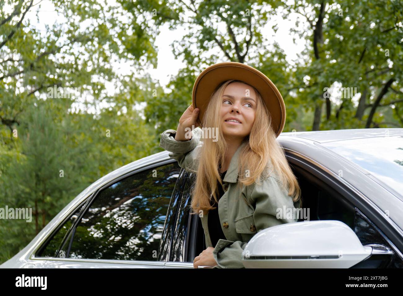 Blonde woman in hat sticking head out of windshield car. Young tourist explore local travel making candid real moments. True emotions expressions of getting away and refresh relax on open clean air Stock Photo