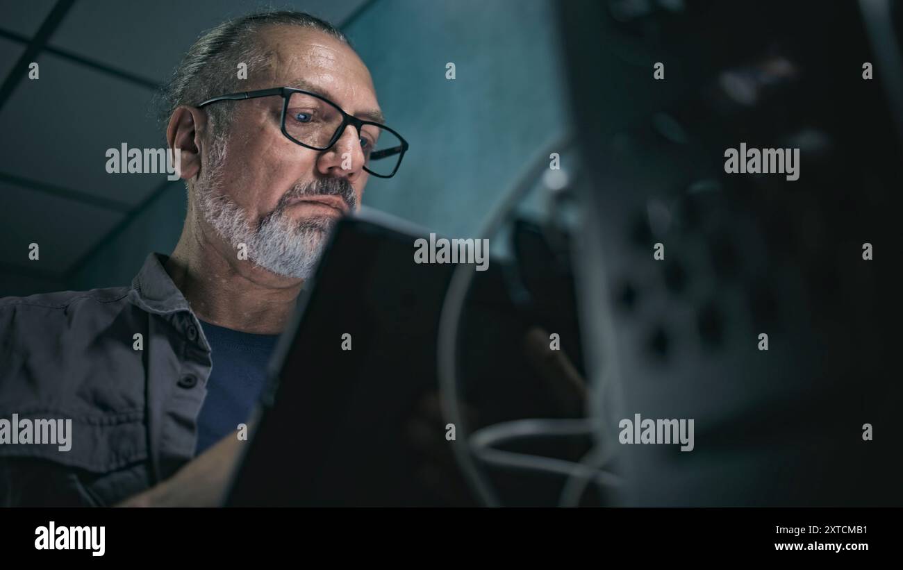 Close up of mature specialist inserting cable into server rack. Network administrator works in server room using tablet computer. Operation of information systems and networks. Data storage center. Stock Photo
