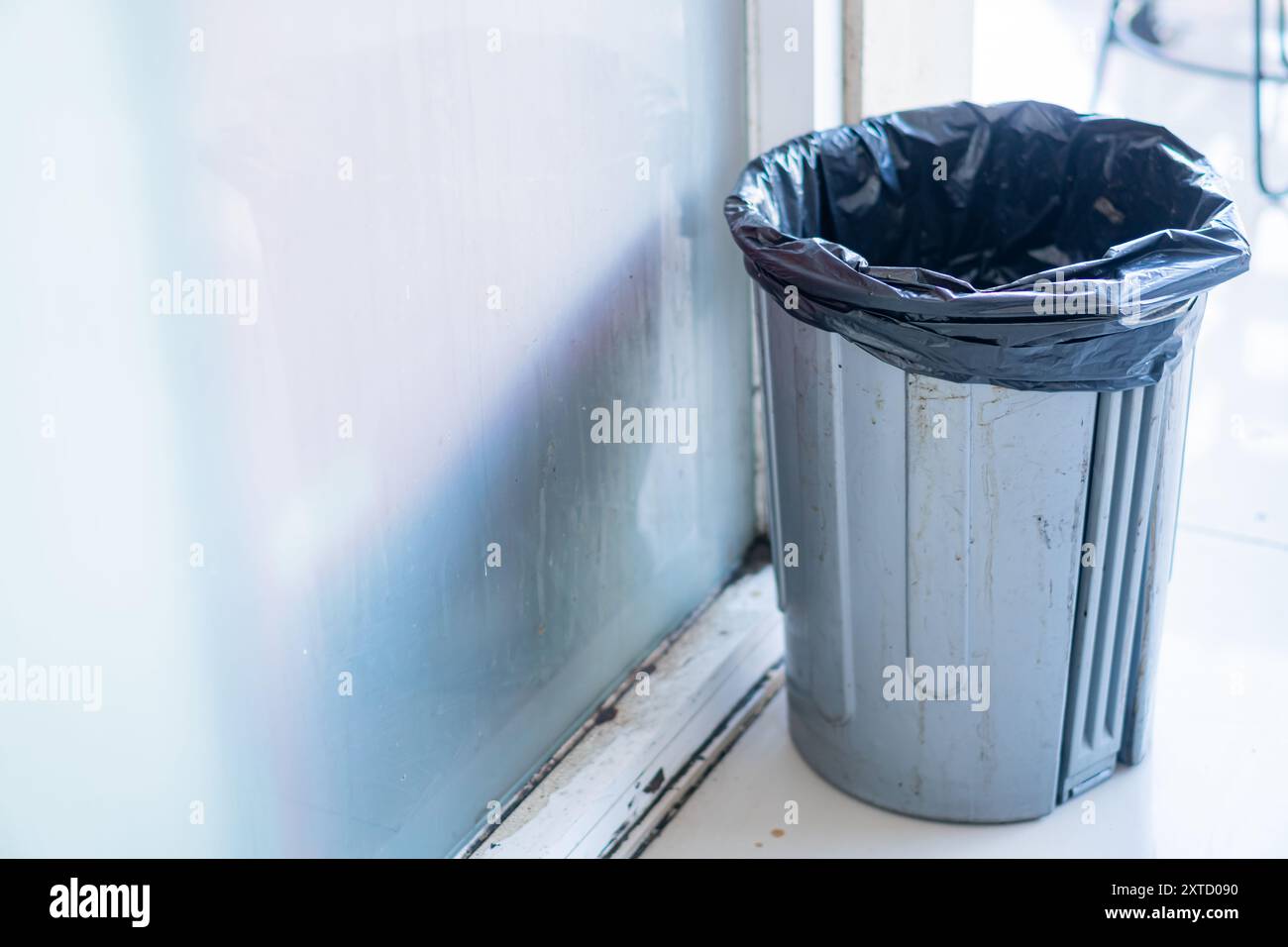 A gray trash can with a black plastic liner, positioned near a window. The can is slightly dirty, indicating use, and the background features a blurre Stock Photo