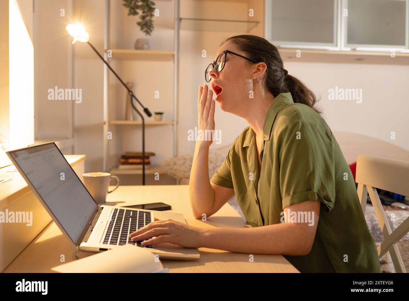 Caucasian young woman is sitting at her desk with a laptop and yawning. Fatigue due to overworking. The student falls asleep near the computer. Online Stock Photo