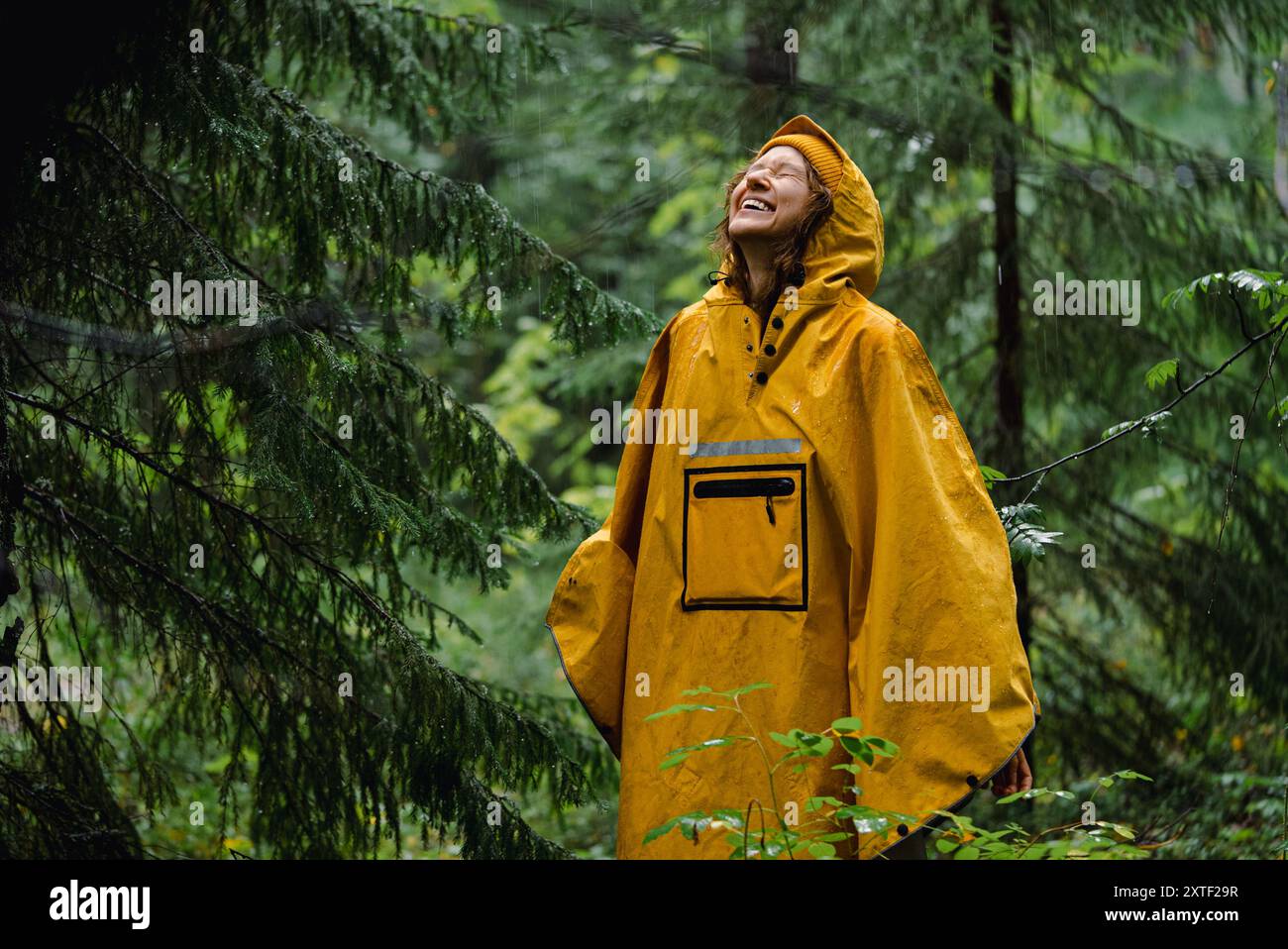 Joyful woman in yellow raincoat embraces rain while standing in lush ...
