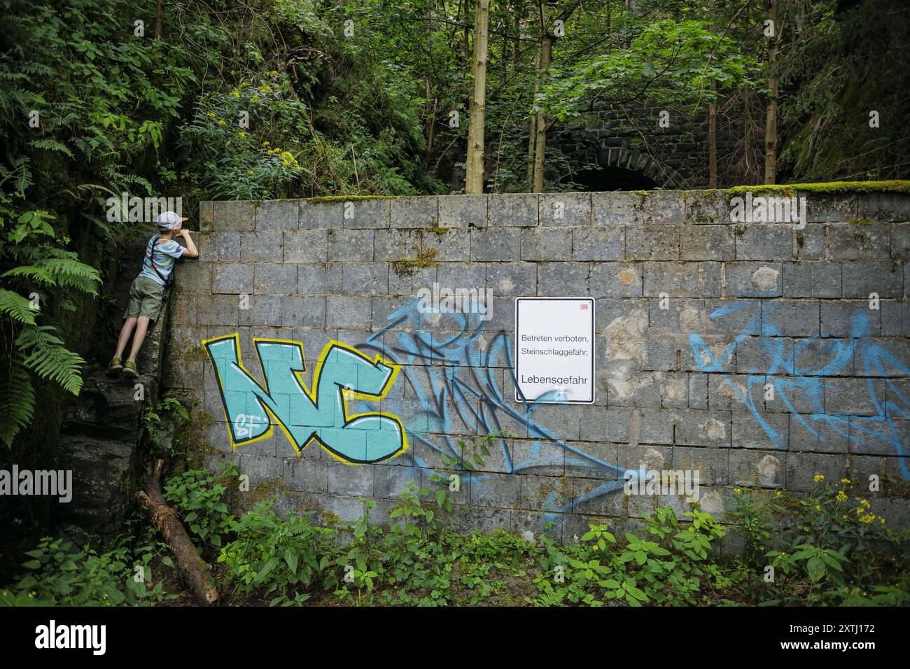 Symbolfoto zum Thema Neugier bei Kindern. Ein Junge steht auf einem Stein und schaut ueber eine Mauer an der ein Hinweisschild mit der Aufschrift ??? Stock Photo