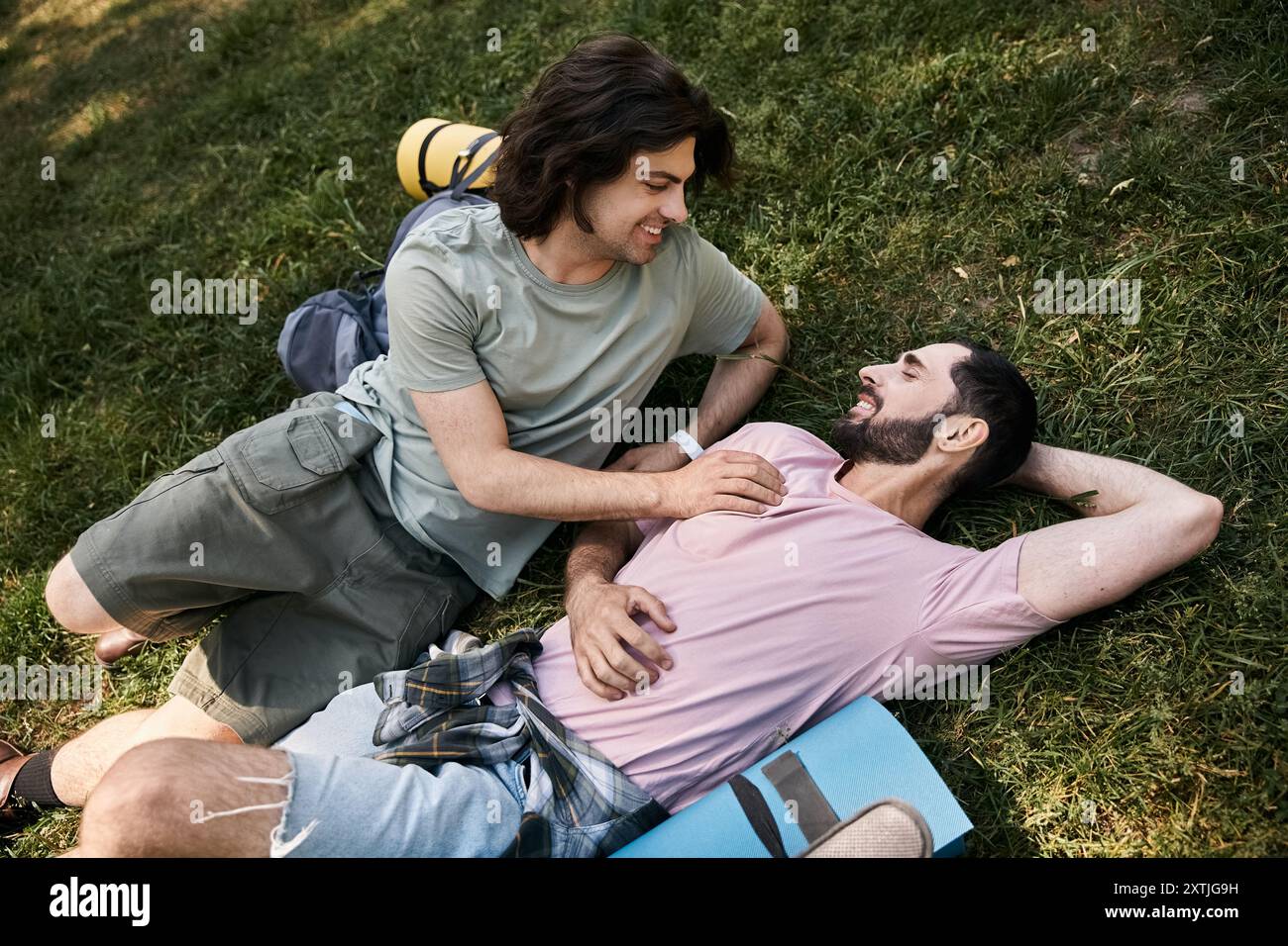 Two young men, a gay couple, enjoy a summer hike, laying on the grass and smiling at each other. Stock Photo