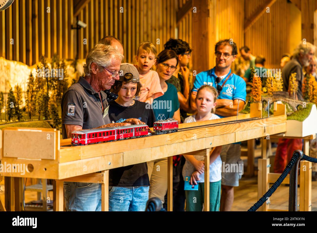A family with several children watches an older man run a red model train along a wooden layout in the model-railway workshop of the Albula Railway Museum, Bergün Filisur, Grisons. The Albula Railway Museum opened on 2 June 2012 in a former arsenal building erected 1917–1919 opposite Bergün station and tells the history of the Rhaetian Railway in the Albula valley. Spuenda da Latsch, Bergün Filisur, Grisons, Switzerland. Stock Photo