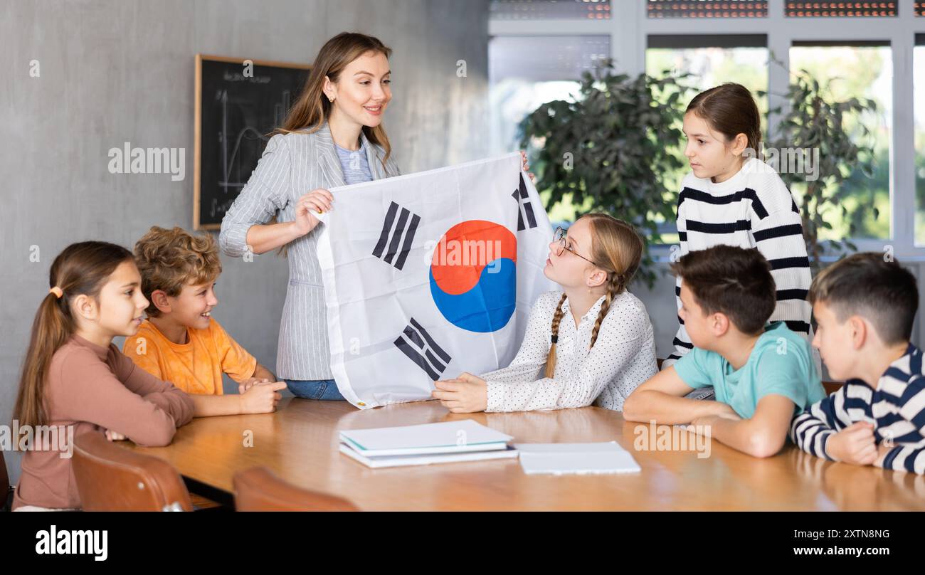 Joyful young female teacher showing flag of South Korea to schoolchildren preteens during history lesson in classroom Stock Photo