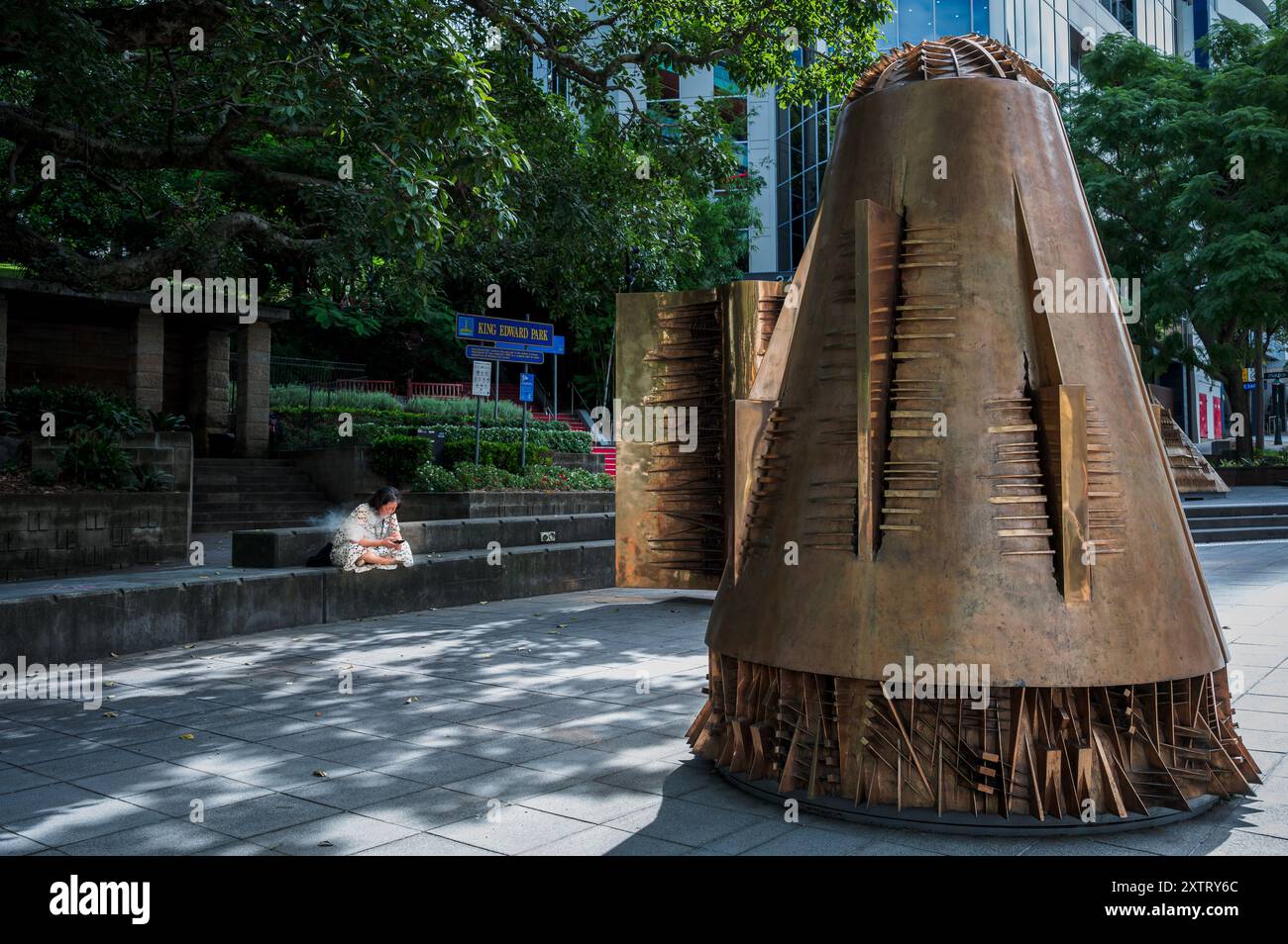 A woman sits cross-legged vaping and looking at her phone in a public space enjoying her solitude next to artistic work by Arnaldo Pomodoro. Stock Photo