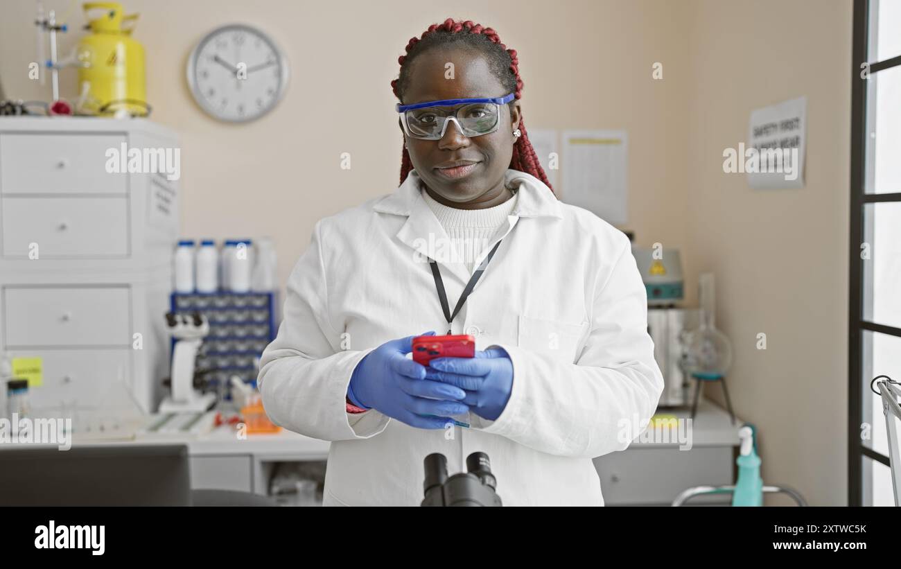 Black woman with braids wearing lab coat and safety glasses uses phone ...