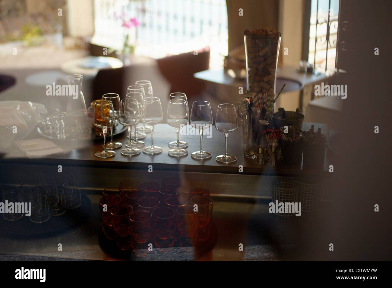 Evening sunlight filters through a window, casting a warm glow over an assortment of empty wine glasses arranged neatly on a bar counter. Stock Photo