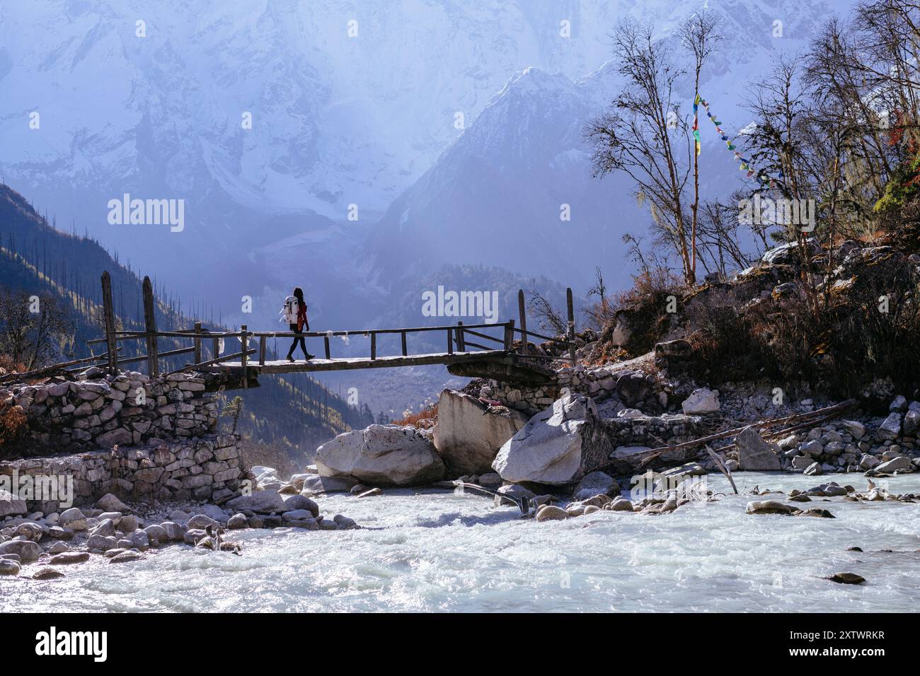 A person crossing a wooden bridge over a mountain river with snow-capped peaks in the background. Stock Photo