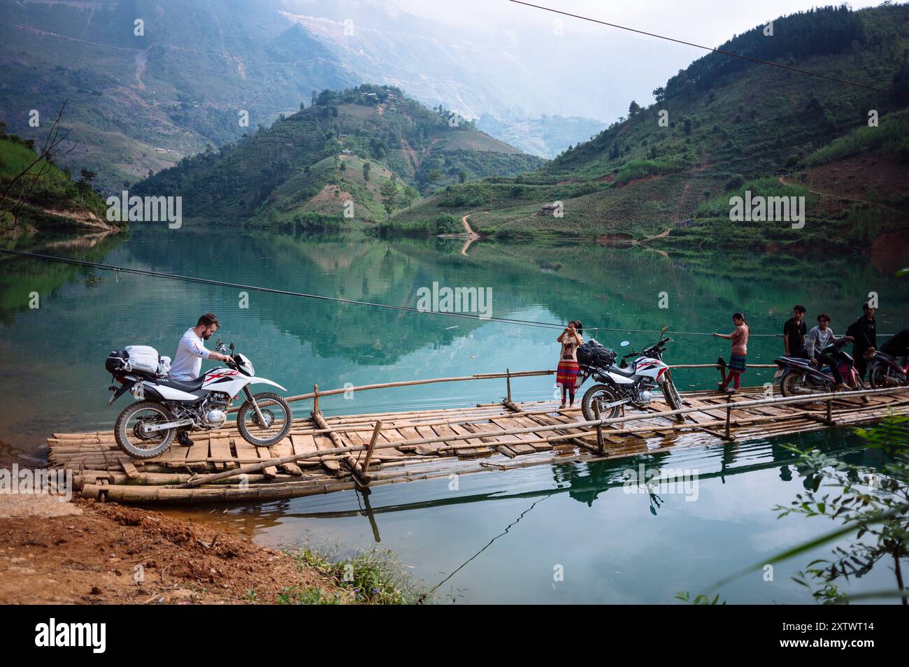 Motorcyclists crossing a rustic wooden bridge over a serene mountain lake with onlookers standing nearby. Stock Photo