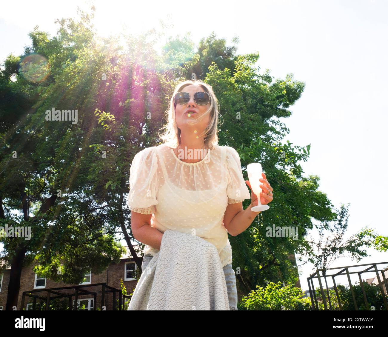 A woman in a white dress and sunglasses stands outdoors holding a drink ...