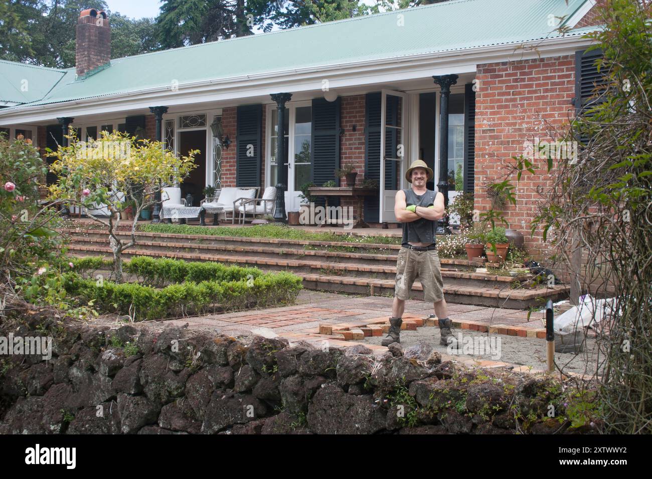 A cheerful home owner stands with arms crossed in front of a beautiful house with an inviting porch and well-kept garden, Auckland, New Zealand Stock Photo