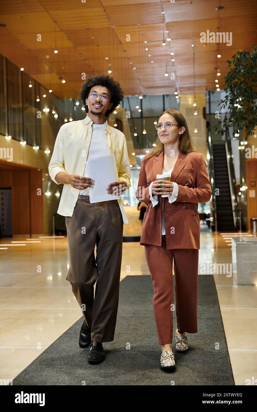 Two colleagues, a man and a woman, walk and chat in an office lobby. Stock Photo