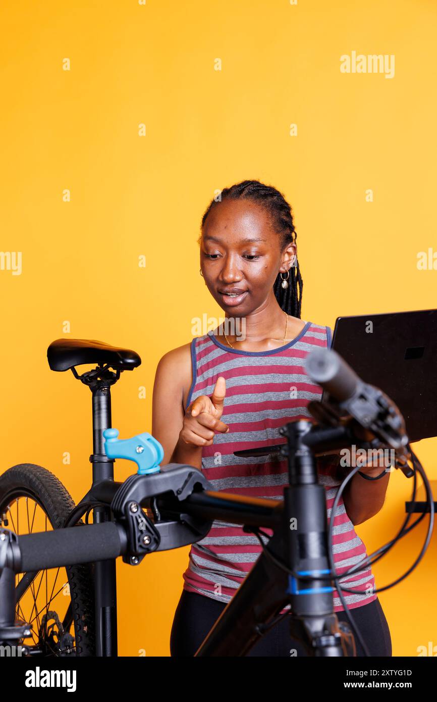 African American woman fixing bike components and using a wireless computer for maintenance and repair. Sports-loving female utilizing laptop in servicing of modern bicycle for leisure cycling. Stock Photo