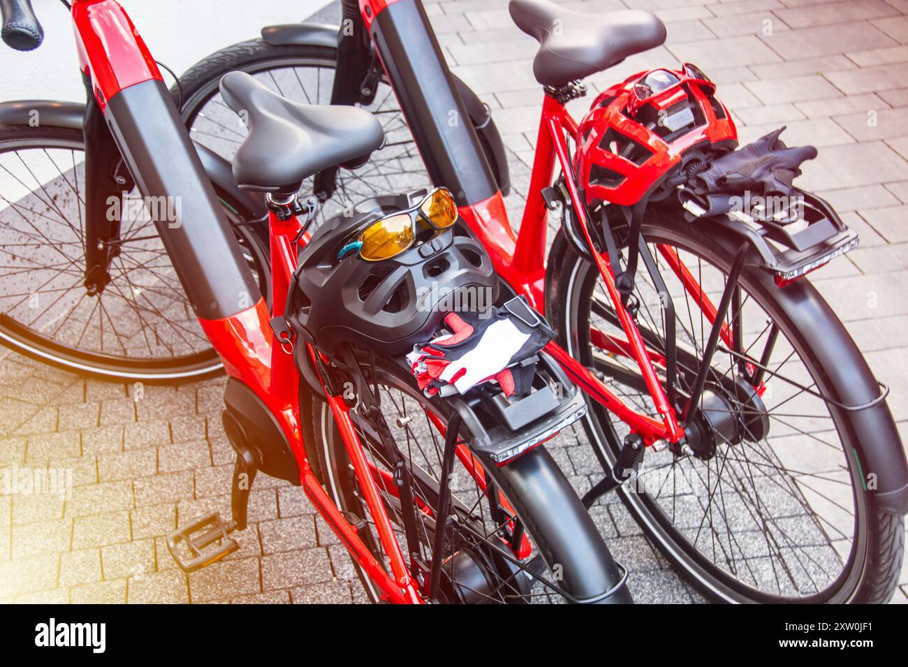 Two vibrant red electric bikes equipped with safety helmets, sunglasses, and gloves rest on a paved surface. Ideal for cyclists seeking convenience, s Stock Photo