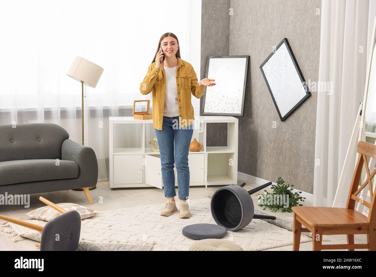 Desperate woman talking on smartphone in messy living room after robbery Stock Photo